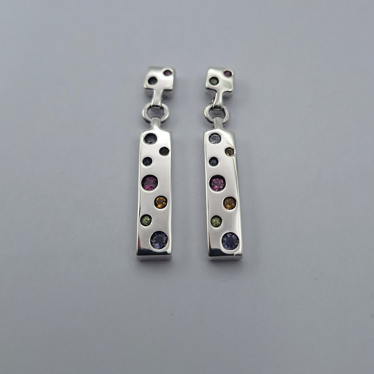 A pair of silver drop earrings with a vertical bar featuring multiple bezel-set round gemstones in various colors, displayed on a neutral gray background.
