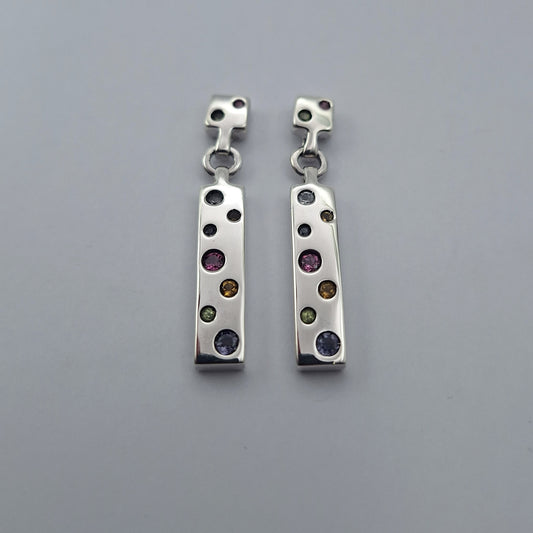 A pair of silver drop earrings with a vertical bar featuring multiple bezel-set round gemstones in various colors, displayed on a neutral gray background.
